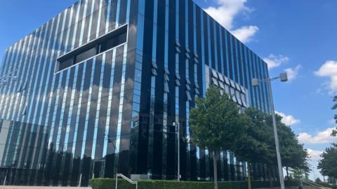 The glass and steel exterior of the Corby Cube building, headquarters of North Northamptonshire Council, on a blue sky day.