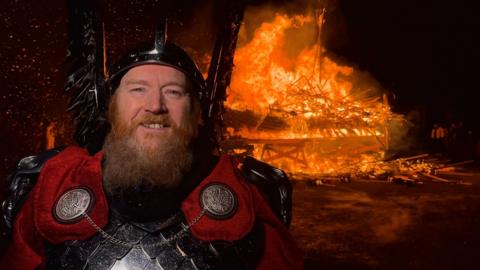 Guizer Jarl Lynden Nicolson wearing Viking armour, standing in front of ship on fire and smiling