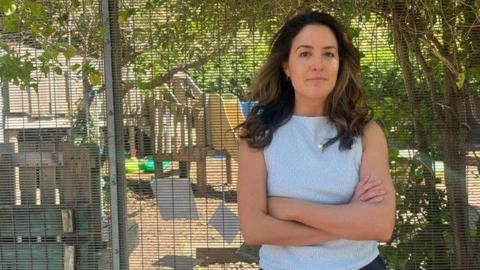 A brown haired woman stands with arms crossed outside a fenced play area at a nursery, with children’s play equipment visible behind