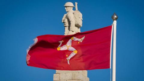 The stone carved World War One soldier that stands on the top of the Douglas war memorial with a red Manx flag flying across it, set against a clear blue sky.
