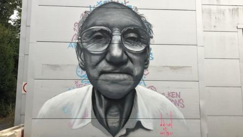 A mural of an elderly man, who has on glasses, looking straight on, painted on a building, with words behind the image and a signature at the bottom. There are trees to the left. 