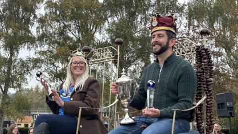 A woman and a man sitting on chairs that say Queen Conker and King Conker across the back. They are both holding silver trophies, wearing crowns and smiling. She has long grey hair and is wearing glasses, a brown coat and blue jeans. He has a dark beard and is wearing a green jumper and blue jeans.