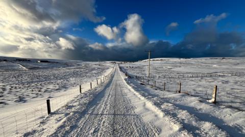 A snow-covered country road stretching to the horizon, under a partially cloudy sky