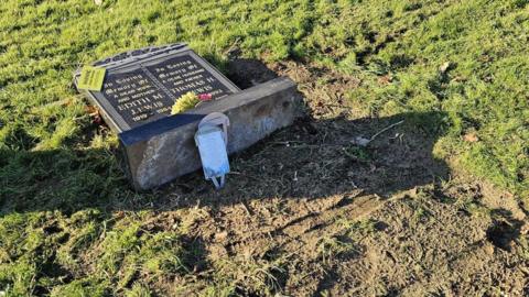 A grave stone knocked over.  On the black stone is two names in gold lettering. There is a yellow warning sign on the top left corner and some flowers at the bottom. Around the stone is grass and muddy soil with tracks in it. 