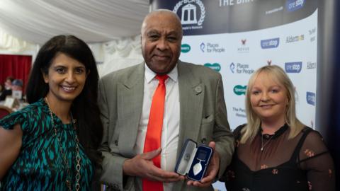 Mick is wearing a light grey suit, white shirt and red tie and standing between Konnie Huq on the left, who is wearing an black and turquoise patterned dress, and Lisa Hannah, who is wearing a sheer black top over a black dress.