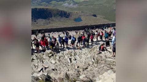A mountain's summit can be seen along with a massive queue of people waiting to reach its peak. The majority of them are looking at the camera. The background is a mixture of grassy and mountainous/rocky landscape.