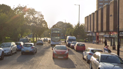 Google maps view of Crow Lane in Henbury, Bristol. Shops can be seen on the right hand side and cars are parked on the side of the street. Other vehicles can be seen driving along. 