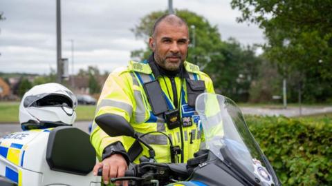 Chris Asante-Ampudah with short dark hair and beard in a police hi-vis jacket sitting on a motorbike