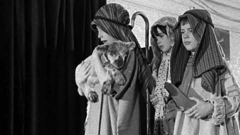 Three children dressed as shepherds. One is carrying a lamb.