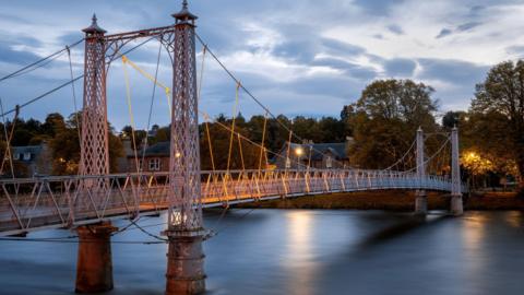 Pictured at night-time, the footbridge is constructed of latticed metalwork and steel cables. The River Ness flows below the crossing. Trees line a bank of the river.