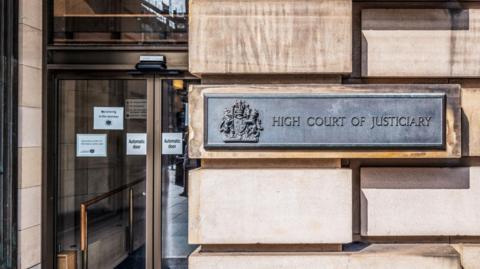 An exterior view of the High Court in Edinburgh showing its glass front door and a sign that says "High Court of Justiciary" on a sandstone wall.