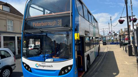 A Blue and white Stagecoach bus at a bus stop in the town of Ramsey,  a silver car is passing the bus and hanging baskets of colourful flowers can be seen on lampposts