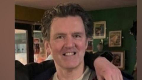John McGarry standing in a pub and smiling. He has swept back hair and is wearing a blue jacket over a light brown jumper and white collared shirt. A man's arm can be seen draped over his shoulder.
