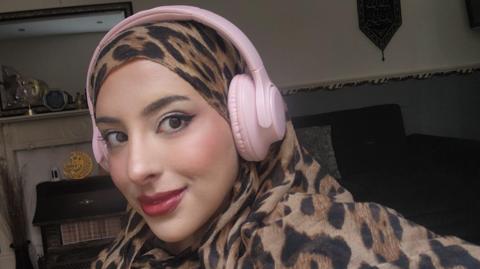 Aisha Sadiqa with a leopard print patterned hijab and pink headphones. She is sitting in a living room with a gas fire and mantlepiece behind her.