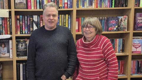 An older man and woman stand side-by-side in front of a bookshelf filled with colourful books. The man has short grey hair 