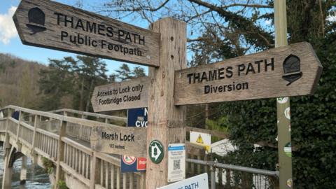 A wooden sign by a bridge over a river, indicating 'Thames Path public footpath', 'Access to lock only/footbridge closed', 'Marsh Lock' and 'Thames Path diversion'. The Thames Path signs include acorn symbols.
