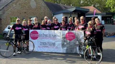 About 20 cyclists all wearing black cycling outfits stand with their bikes and hold up a large white banner for the Worcestershire Breast Unit Haven. The tag line "everybody knows somebody" is also printed on the banner with a phone number and website for the service.