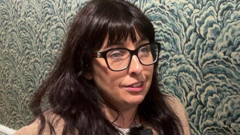 Amy with long brown hair and glasses on in front of a patterned wall.