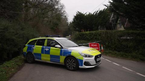 A Devon and Cornwall Police car is parked across a road. Behind it is a red 'road closed' sign, a cone and police tape. It is parked on a rural street with hedges and overhanging trees.