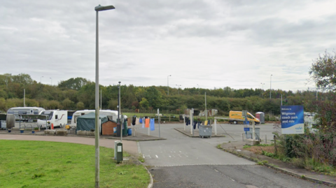 A wide view of Wigmore Coach Park showing caravans, temporary structures, washing lines and a sign at the site entrance.