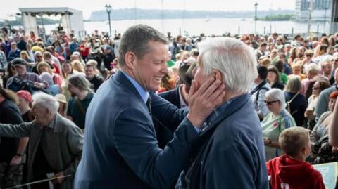 Two men, one of whom is Rhun ap Iorwerth, speak in front of a big crowd of people