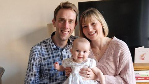 A blonde woman in a pink jumper sits next to a blonde man wearing a checked shirt. She is holding a baby in a white dress.