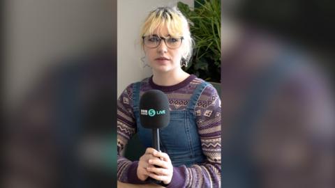 A teenage girl holds a 5 Live mic. She has short blonde hair and wears a purple patterned jumper and denim dungarees