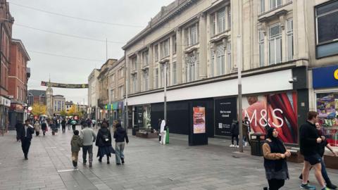 Shoppers wander past a large vacant unit once home to Marks and Spencer on a pedestrianised street, with the city's famous Clock Tower in view down the road