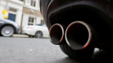 An exhaust pipe on the back of a car. A wheel can be seen, with two cars out of focus in the background