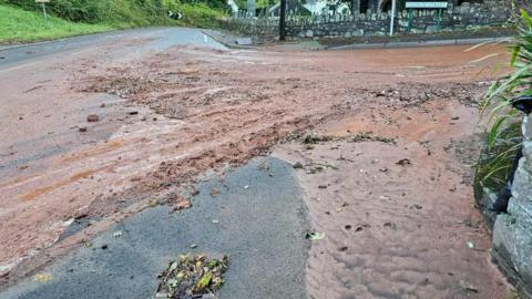 A flooded road mixed with mud besides a pub in Totnes. The water is brown with mud and there is a brick wall on the opposite side of the junction.