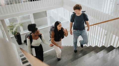 Three students walking up a flight of stairs, wearing backpacks.