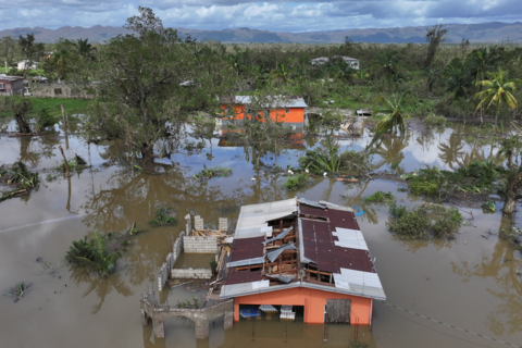 Drone view of flooding after Hurricane Melissa made landfall in St Elizabeth, Jamaica, October 29, 2025. Two orange-painted houses are partly submerged in water and large sections of the roofs are missing. In the distance are palm trees and mountains.