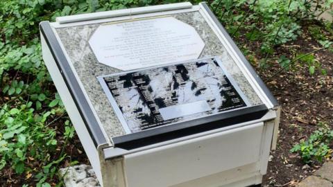 The damaged plaque in a memorial garden. It is a stone plinth with a granite top, where the names of children killed in the Blitz are displayed in a black plaque. A piece of grey duct tape covers up a scratch.