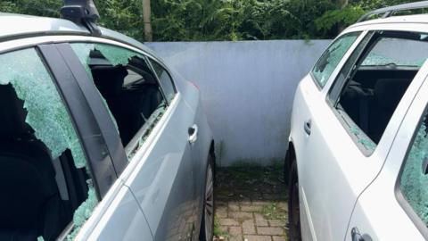 Two cars parked next to each other at a car garage site. Both vehicles have had their side windows smashed.