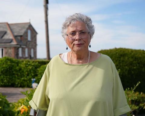 carole wyatt, a woman with short grey hair, wire frame glasses and a pale green tshirt, stands in front of a hedge and a house