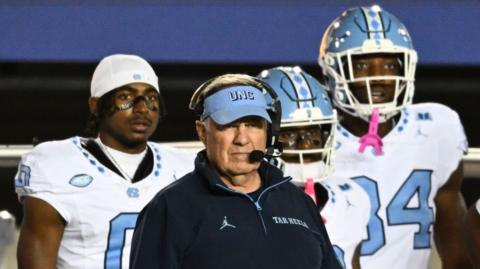Bill Belichick wears a headset as he stands on the sideline during a game. Three football players in white and light blue uniforms stand behind him.