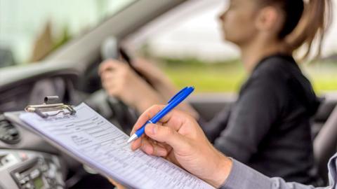 A driving instructor holds a pen to a clipboard in a car whilst a young woman takes her test in the driving seat