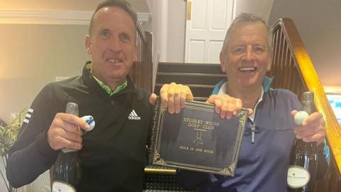 Steve Wilmshurst and Liam Nairn smile and hold up the Studley Wood Golf Club hole-in-one book. They each have a bottle of champagne and a golf ball.