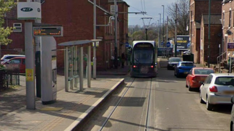 Tram stop is pictured with a green tram leaving the station and a row of cars on the right