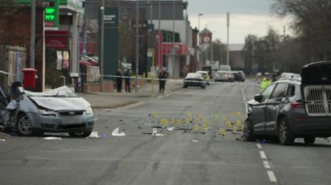 A grey-blue Ford Focus lies across a road with its back smashed and much of its roof sliced off. A dark-coloured police car, with its front damaged and back gate open, stands opposite. Debris is scattered across the cordoned-off section of road. Yellow crash-scene investigation markers have been put in place on the road.