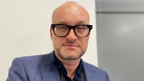 A bald man wearing a blue jackey and black glasses looks at the camera, against a white background.