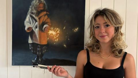 A young artist with blonde hair stood next to her art - a painting of a young girl in a coat holding a sparkler.