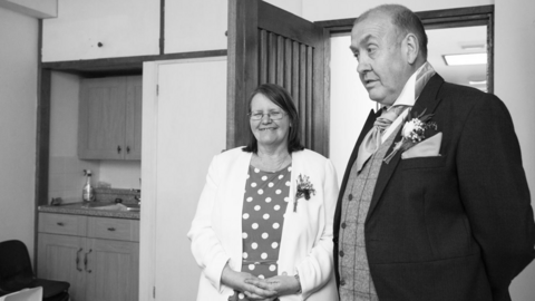 A black and white photo of Linda and Bob dressed in smart clothes as wedding guests in a reception room with a sink and cupboards behind them