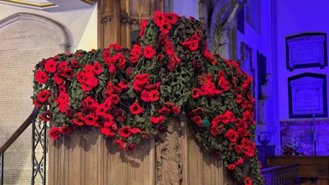 A poppy drape, hanging on a church pulipt. It features knitted red poppies attached to green cargo netting. The building behind is illuminated with a blue light.