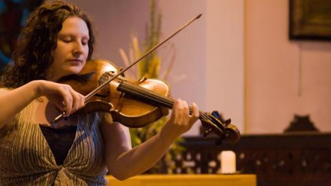 Laura Rowles, a woman with dark curly hair, looking down at the strings as she plays the violin.
