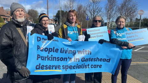A group of people wearing winter outwear and blue t-shirts reading Parkinson's UK stand together. They are standing on a street holding a blue banner which reads - people living with Parkinson's in Gateshead deserve specialist care.
