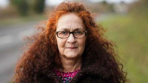 A woman with long curly auburn hair wearing a brown coat and wearing glasses is stood facing the camera with a blurred background