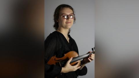 A professional photo of Esther holding her violin under her arm and smiling to camera. She is wearing glasses and a black formal blouse. There is a neutral light grey background colour.