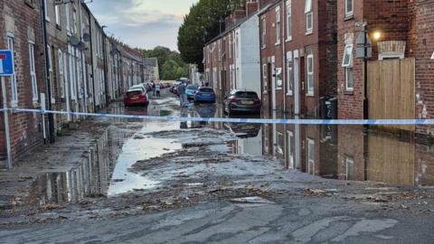 Police tape cordening off a partially flooded road with terraced houses on each side along with a number of cars.