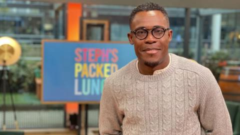 Peter on the set of British daytime television programme Steph's Packed Lunch. Peter is smiling at the camera and is wearing a smart jumper with a shirt underneath. He looks relaxed and content. 